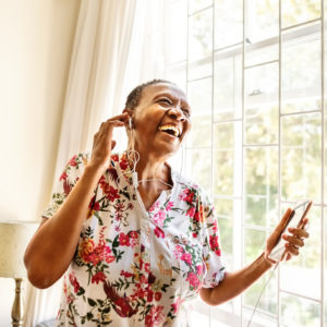 Cheerful senior female standing by window enjoying listening to music on earphones